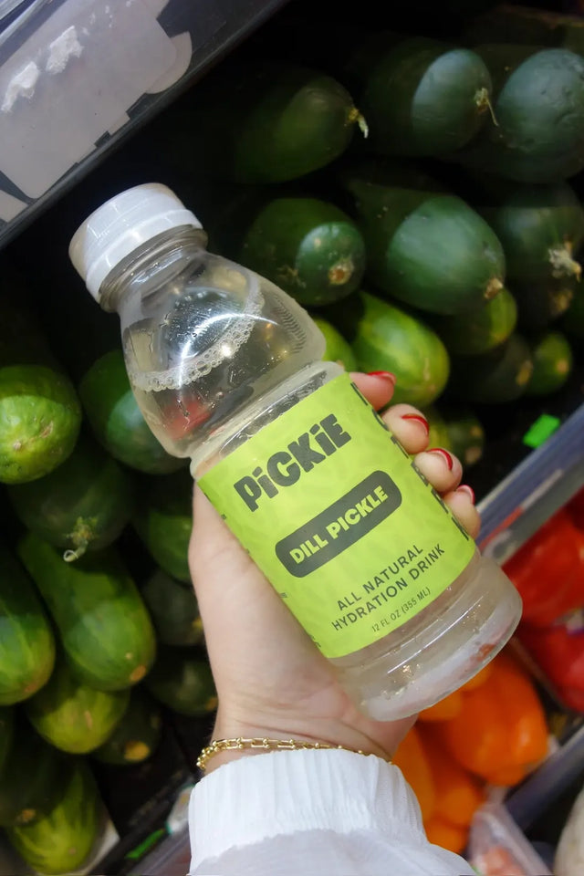 Hand holding PiCKiE Dill Pickle in front of fresh cucumbers