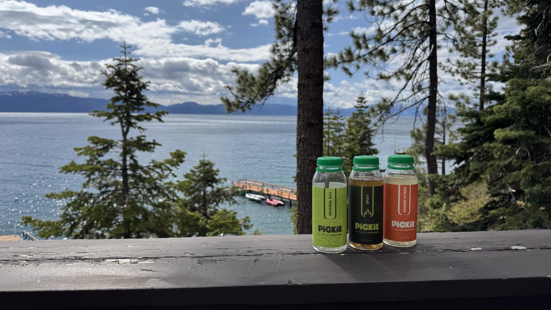 PiCKiE Original Dill, Spicy, and Orange Citrus pickle juice bottles lined up on a railing with a scenic lake, trees, and blue sky in the background.