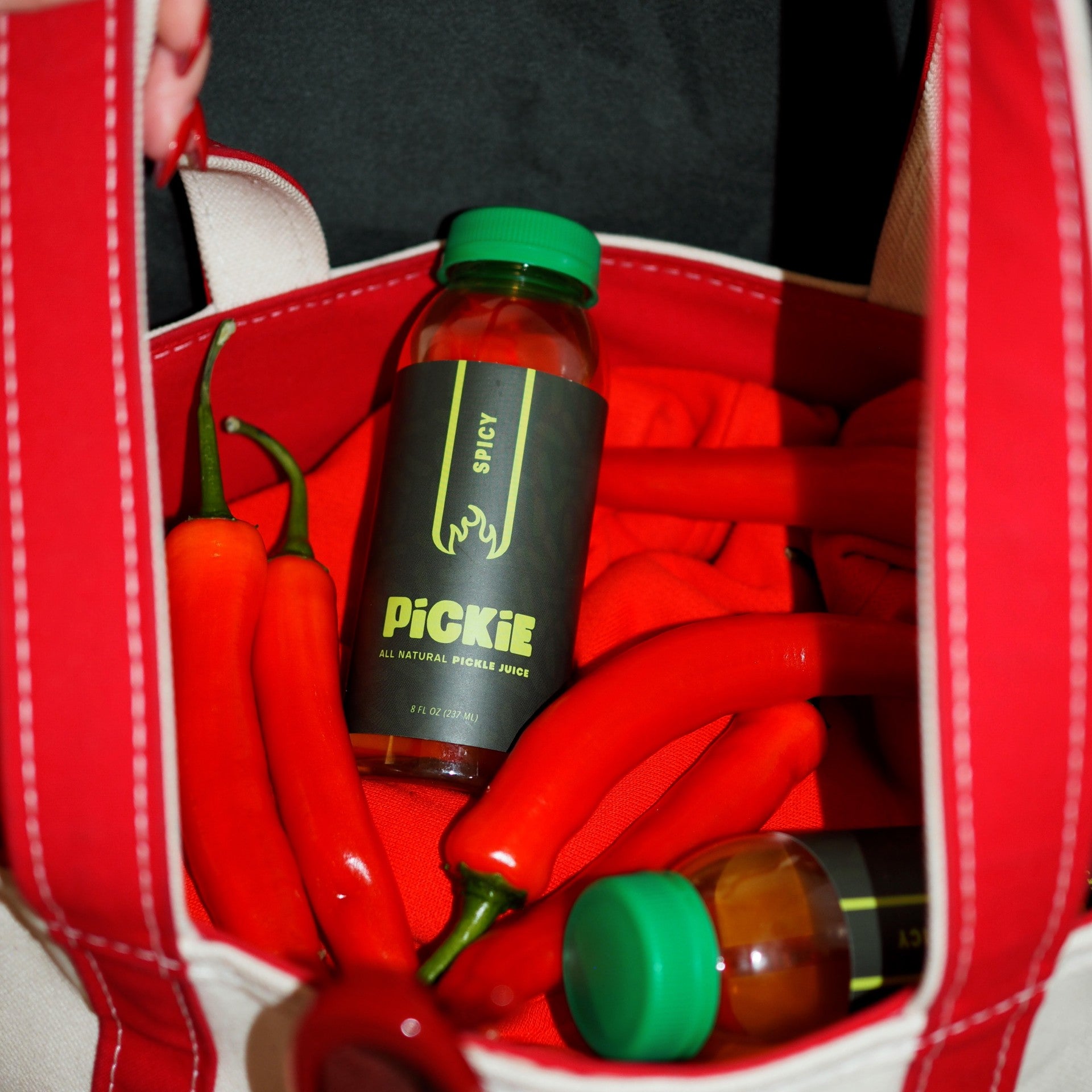 PiCKiE Spicy pickle juice bottle in a bag alongside glossy red peppers, highlighting its bold, pepper-infused flavor and natural heat.