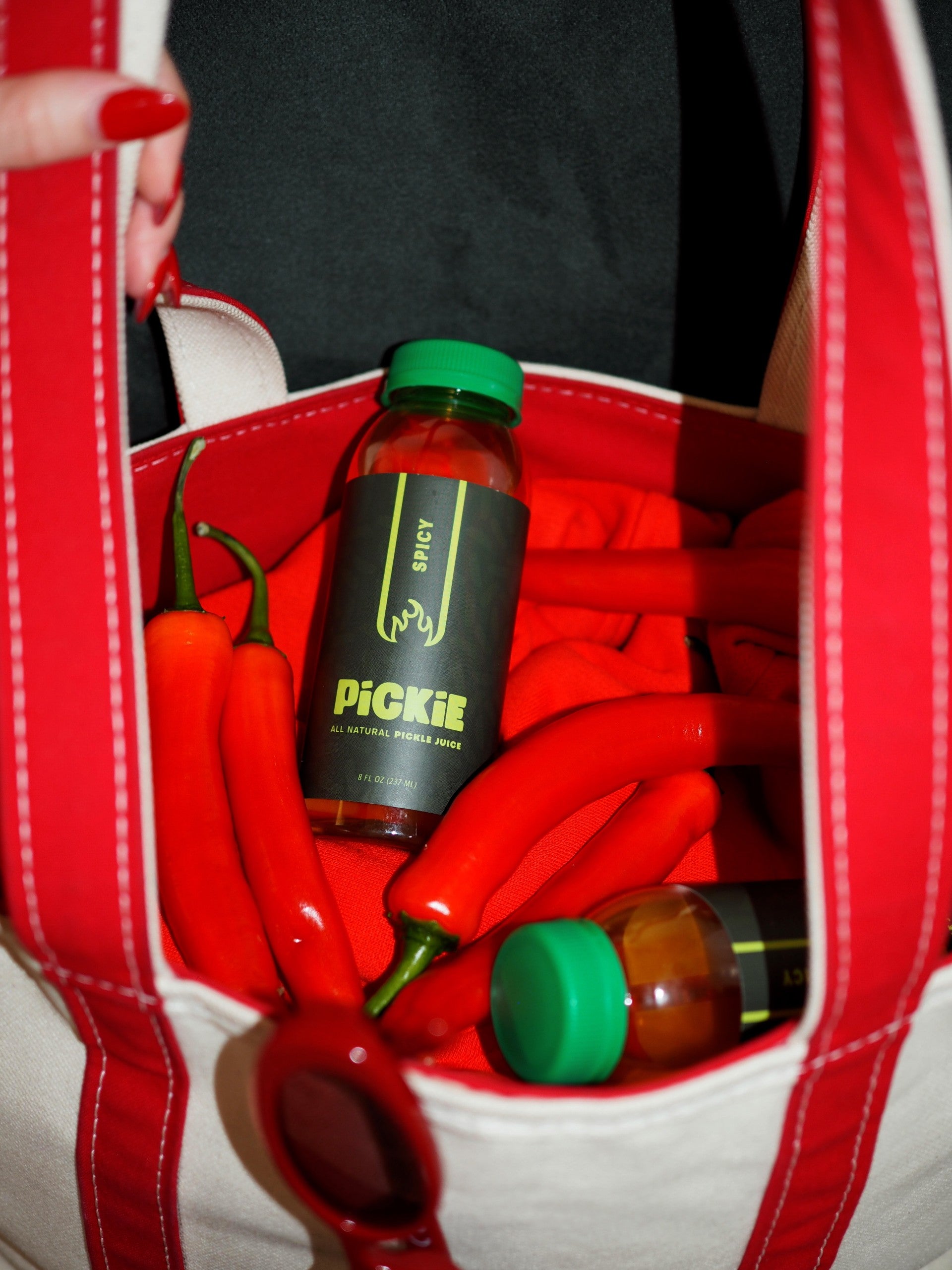 PiCKiE Spicy pickle juice bottle in a bag alongside glossy red peppers, highlighting its bold, pepper-infused flavor and natural heat.