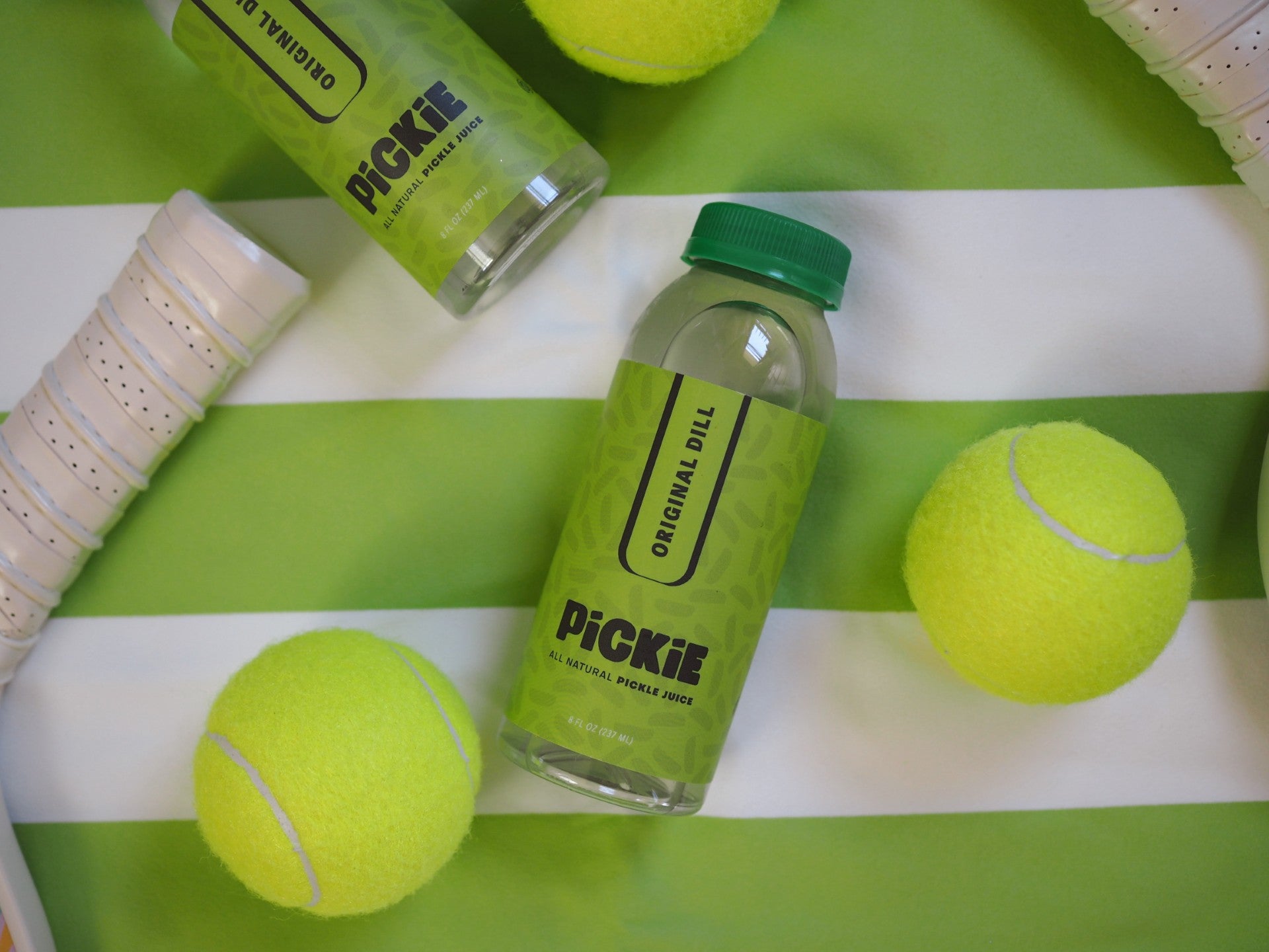 PiCKiE Original Dill pickle juice bottle placed alongside tennis balls and tennis rackets, highlighting its connection to hydration and athletics.