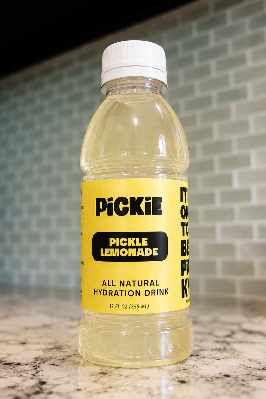 A bottle of Pickle Lemonade all natural hydration drink, featuring a bright yellow label and white cap, set on a granite countertop with a tiled backsplash in the background.