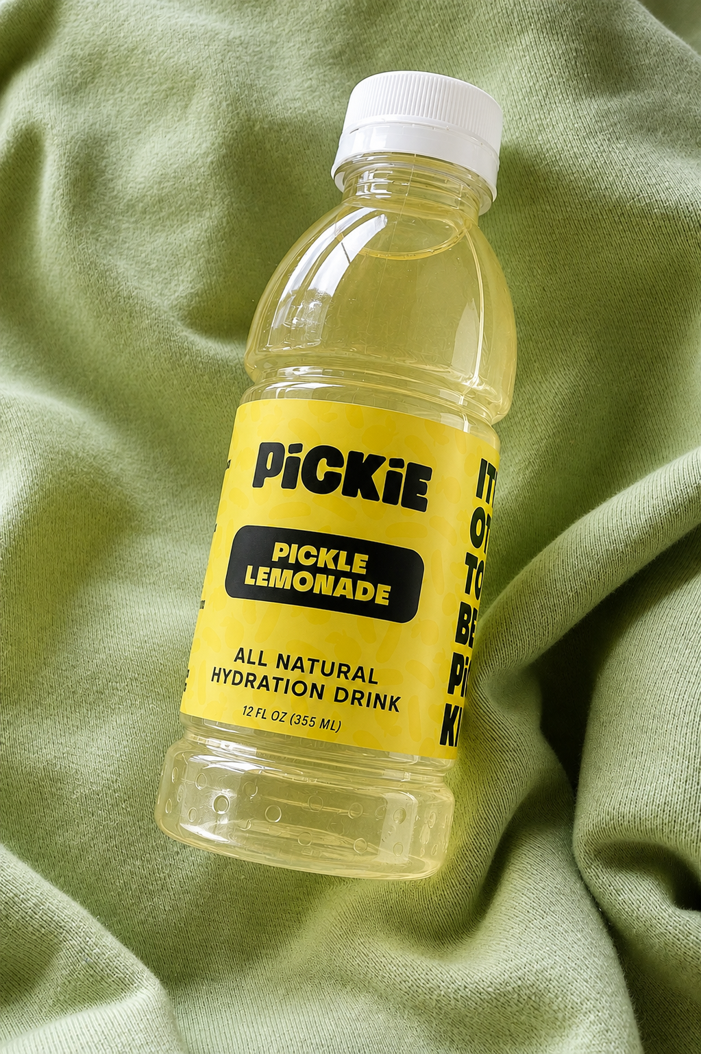 A 12 fl oz bottle of PickKie Pickle Lemonade all natural hydration drink, featuring a bright yellow label and white cap, resting on a textured sage green fabric background.