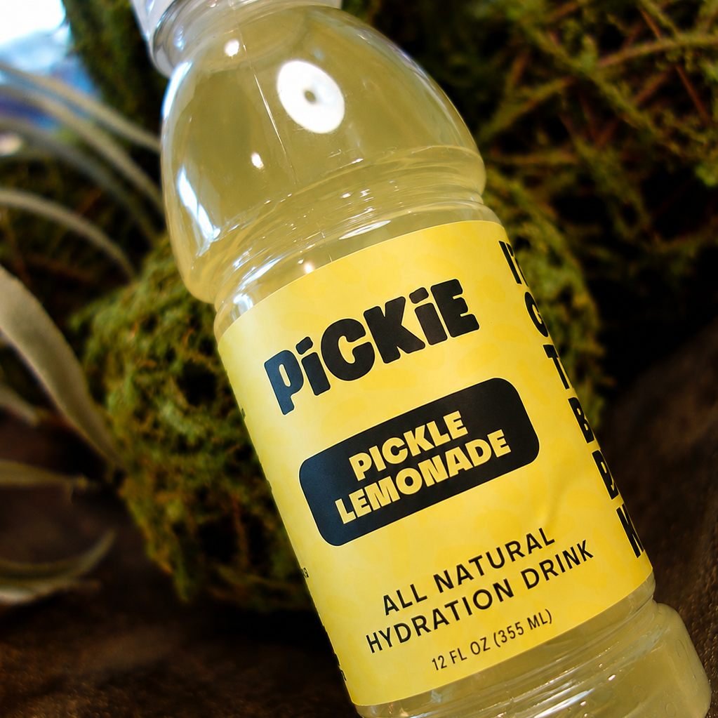 A bottle of PiCKiE Pickle Lemonade all natural hydration drink, featuring a bright yellow label and clear plastic bottle showing the pale yellow liquid inside, set against a background of green moss.