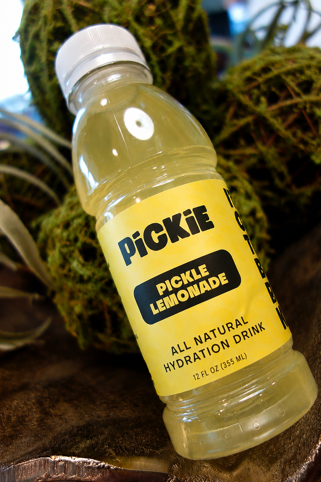 A bottle of PiCKiE Pickle Lemonade all natural hydration drink, featuring a bright yellow label and clear plastic bottle showing the pale yellow liquid inside, set against a background of green moss.