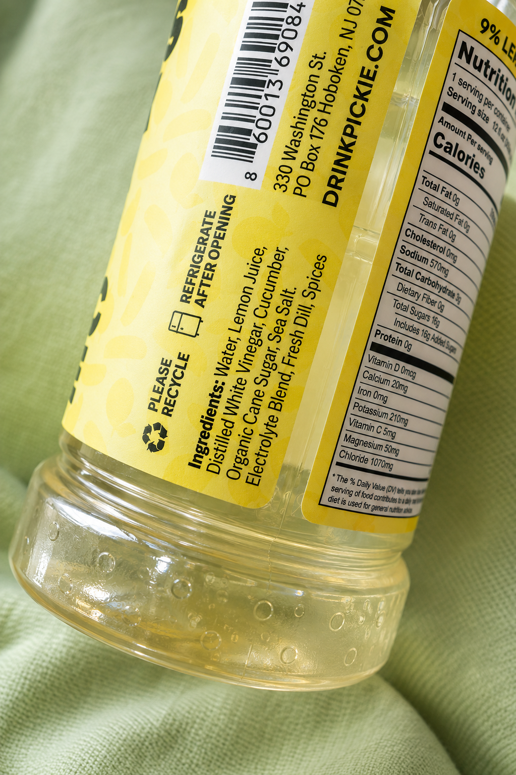 A close-up of a clear plastic bottle of electrolyte drink mix with a yellow label, showing the back of the package including barcode, ingredients, and nutrition facts, resting on a light green textured fabric.