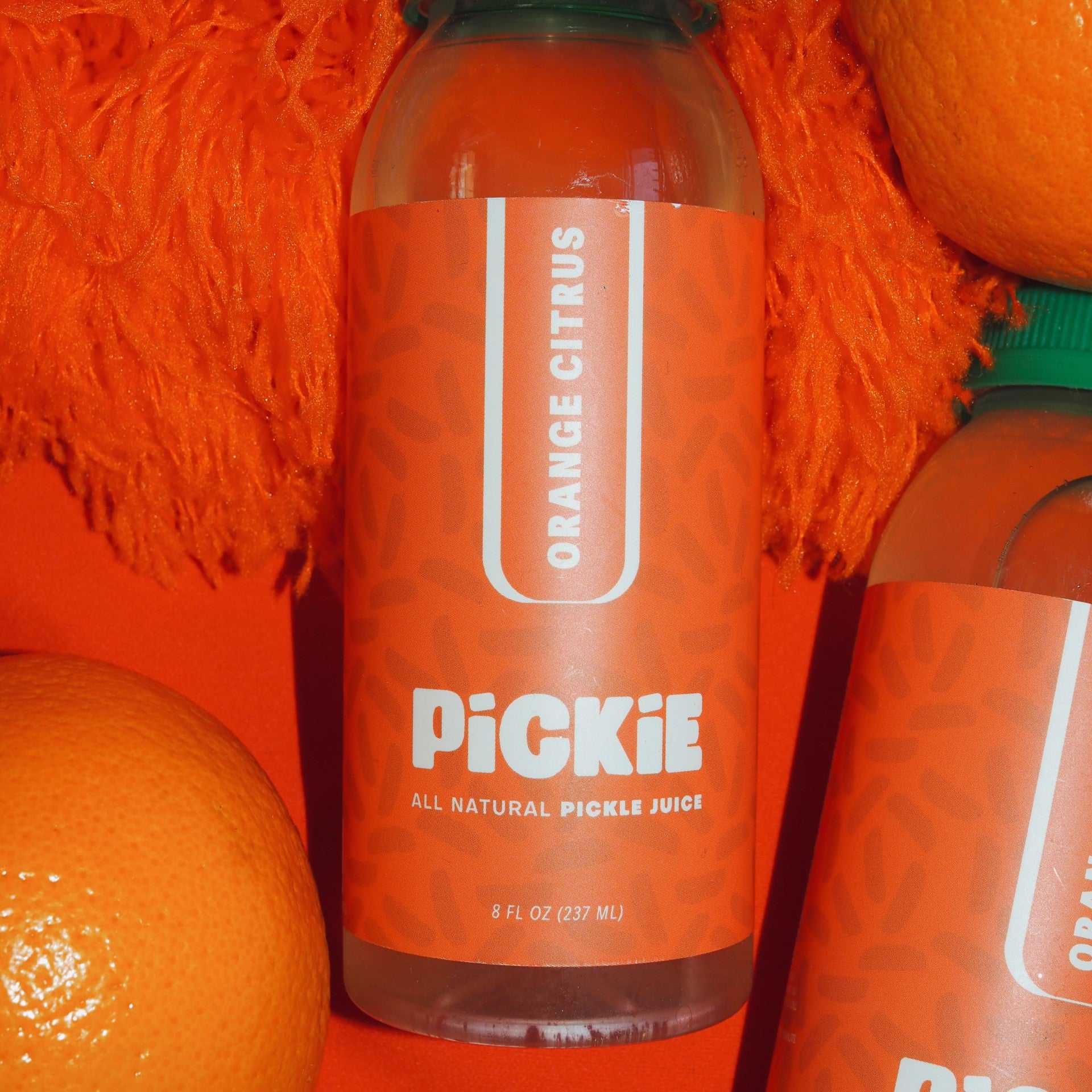 PiCKiE Orange Citrus pickle juice bottle surrounded by fresh oranges on a vibrant, fizzy orange background, emphasizing its citrus flavor and light and refreshing flavor. 