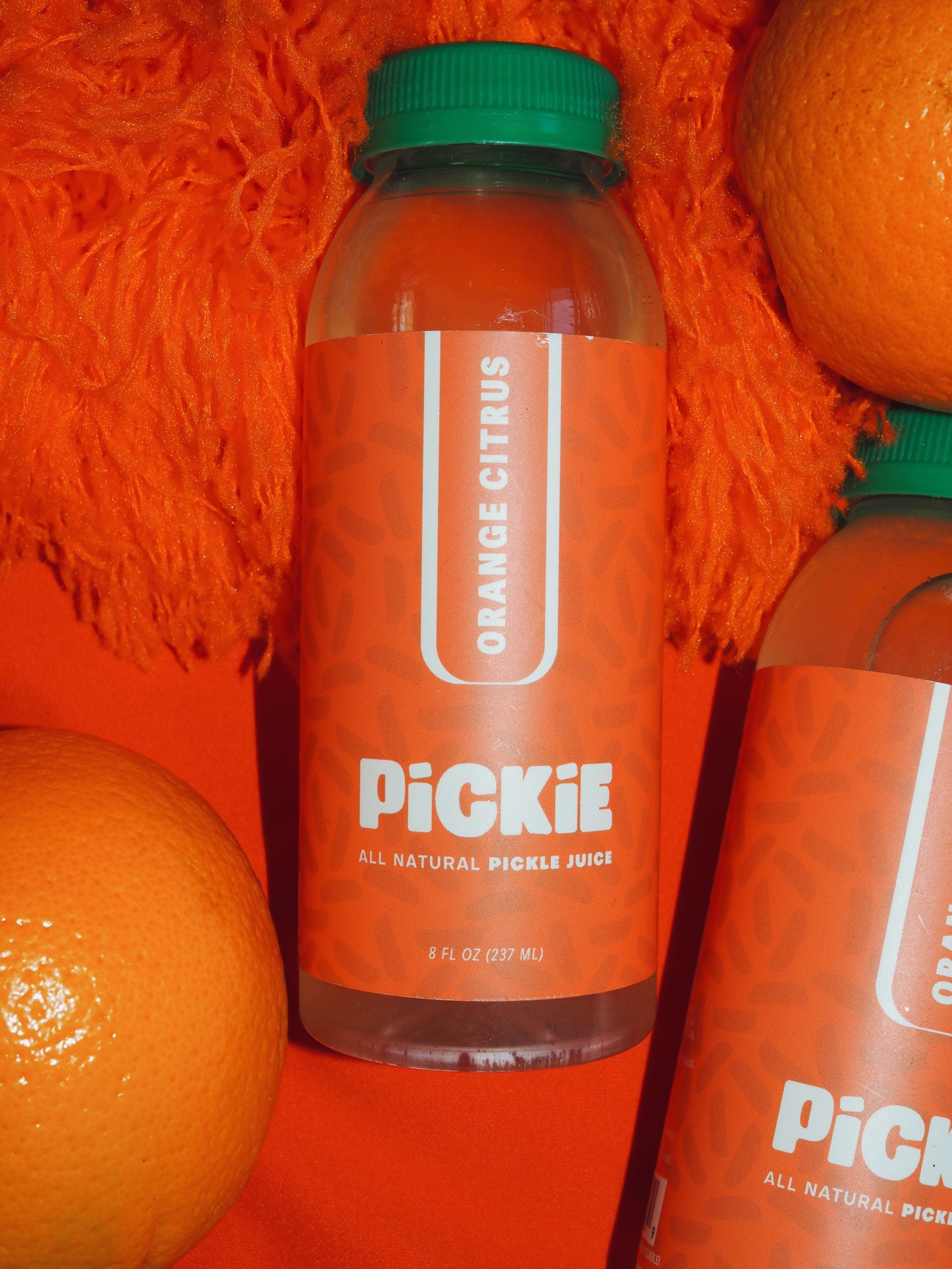 PiCKiE Orange Citrus pickle juice bottle surrounded by fresh oranges on a vibrant, fizzy orange background, emphasizing its citrus flavor and light and refreshing flavor. 