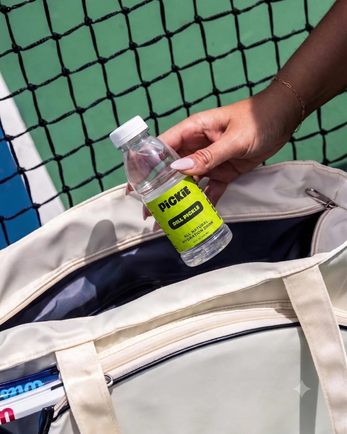 A hand holding the real PiCKiE Dill Pickle bottle above a sports bag beside a court